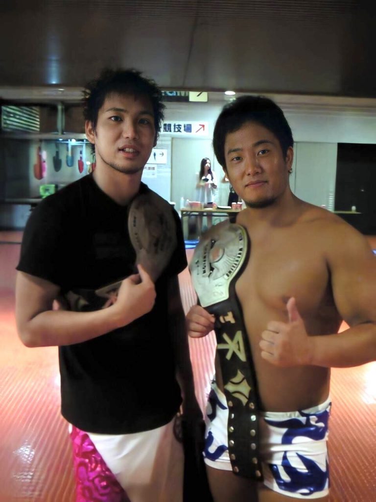 Kotoge Atsushi and Harada Daisuke, standing side by side, both in wrestling gear. Over each of their shoulders is an osaka pro wrestling tag belt. 