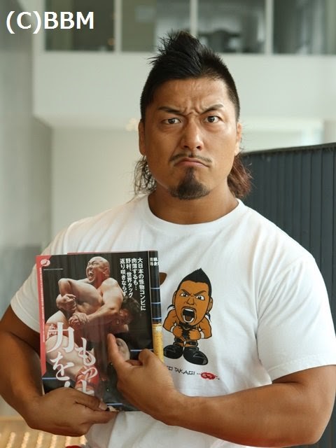 Shingo posing and grimacing, holding a copy of weekly pro wrestling