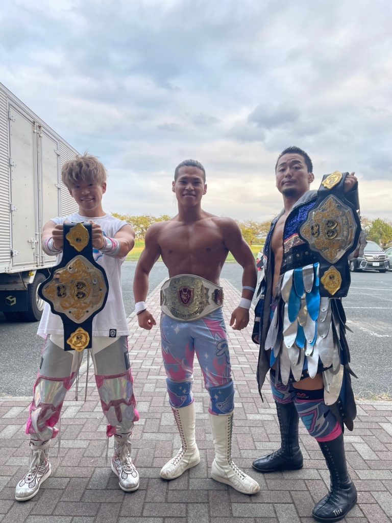 D'Courage (Dia, Ryoya, Yoshioka) stand in a trio, holding up their belts. 