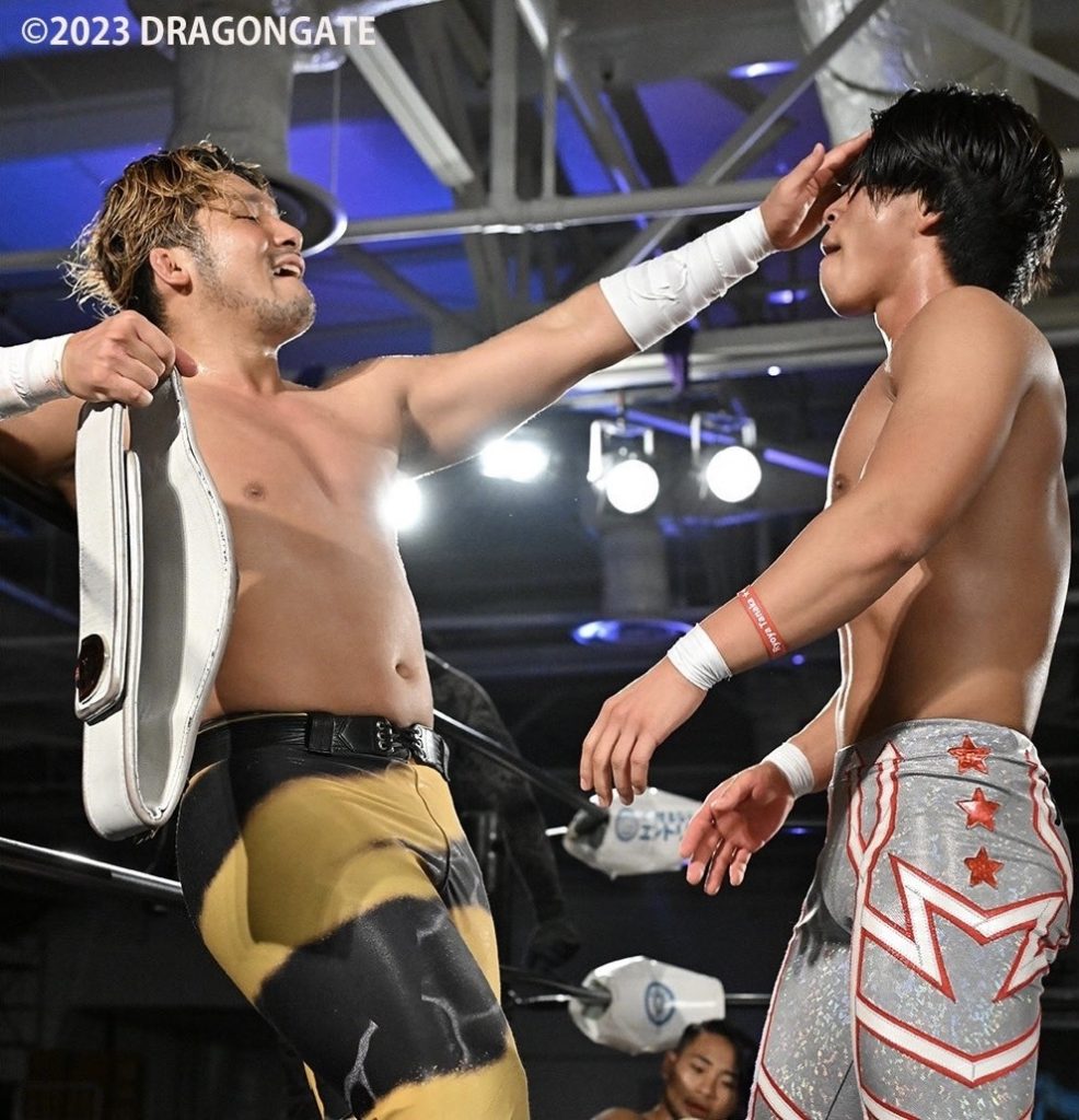 ISHIN leans against the rope, holding the Bravegate belt in one and and pushing away Ryoya in the face with the other. ISHIN looks smug.