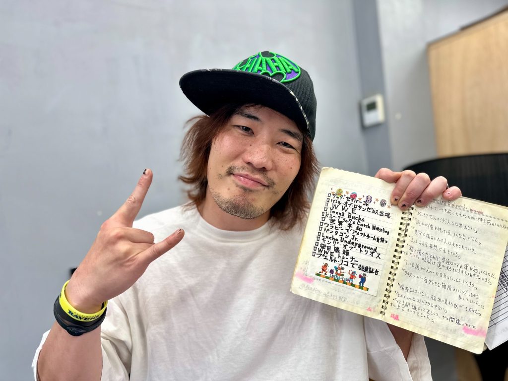 Yano, wearing a cap and t-shirt, holding up a tattered notebook to the camera. On one page is a check list in English and Japanese, seemingly of people or places to wrestle. On the other is notes in Japanese under the English heading 'Charles Bukowski beatnik'