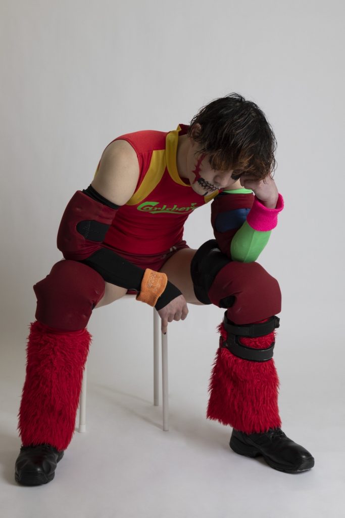 Yano as one of his many personas, KEITA in THE House. Keita is in full gear, sat on a low stool, kneeling forward. His head rests on one fist, his other arm resting on his knee, in a thinking pose.